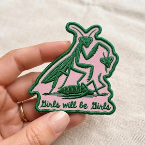 May include: An embroidered patch featuring a pink background with a dark green outline and a dark green illustration of two praying mantises. The text Girls will be Girls is embroidered in dark green cursive script at the bottom of the patch.