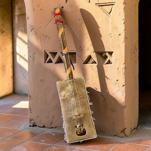 May include: A stringed musical instrument featuring a rectangular brass body with a circular sound hole and a long wooden neck. The neck is wrapped with yellow and blue ribbons and red cord. The instrument has thin metal strings and a bridge.