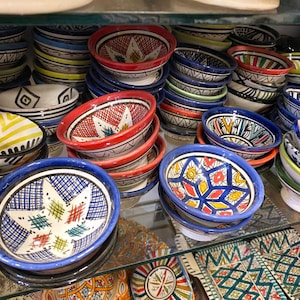 Puede incluir: Una colección de pequeños cuencos de cerámica redondos apilados en estantes de cristal. Cada cuenco presenta patrones geométricos pintados a mano en colores vibrantes, incluyendo rojo, azul, amarillo y verde sobre un fondo blanco. Los diseños incluyen estrellas, rombos y motivos de cuadrícula.