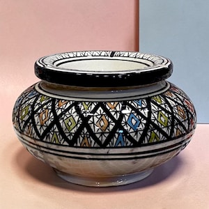 Pode incluir: Um cinzeiro de cerâmica redondo com uma borda preta larga e uma base branca. O exterior é decorado com um padrão de treliça de diamantes pretos repetitivos, com pequenos detalhes de tinta laranja, azul e verde preenchendo cada forma.