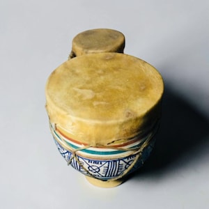 May include: A double-headed drum featuring two circular tan skin drumheads secured with thin cord lacing. The drum body is decorated with horizontal bands of red, green, and blue, with a blue geometric pattern painted on the lower section.