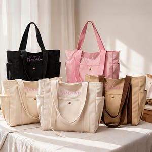 May include: A collection of five canvas tote bags in black, pink, cream, and tan colours. Each bag features two shoulder straps, side pockets, a front pocket with a gold-tone snap button, and a personalised name embroidered in a script font.