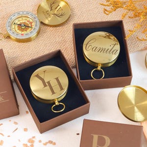 May include: Several gold-coloured metal pocket compasses with hinged lids, some engraved with names like Camila or initials like YH. The compasses feature circular faces with directional markings and are presented in brown boxes with black velvet inserts.