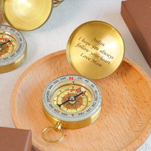 May include: A brass pocket compass with a hinged lid featuring the engraved text: Aidan, I hope you always follow your heart, Love Mom. The compass has a circular face with cardinal directions and degree markings, and a small metal ring.