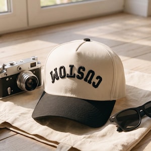 Könnte beinhalten: Eine cremefarbene Baseballkappe mit schwarzem Schirm und dem Wort CUSTOM, das in schwarzen Buchstaben auf der Vorderseite eingestickt ist. Die Kappe liegt neben einer schwarz-silbernen Kamera im Vintage-Stil und einer schwarzen Sonnenbrille auf einer Stofftasche.