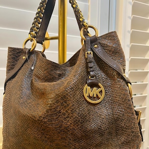 May include: A dark brown hobo bag with a textured snakeskin pattern, featuring gold-tone hardware, a circular MK logo charm, and a braided leather and chain shoulder strap. The bag has a slouchy silhouette with dark brown leather side accents.