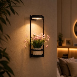 Puede incluir: Una maceta de pared de metal negro con una luz LED circular integrada en la parte superior y un estante en la parte inferior que sostiene una maceta de cerámica blanca con un grupo de flores de color rosa claro. La luz emite un brillo cálido.