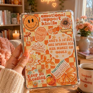 Op de afbeelding: Een doorzichtige beschermhoes voor een e-reader met een oranje en crème geblokte achtergrond, bedekt met diverse stickers, waaronder een smiley, een bloem, een sinaasappelschijfje en de tekst emotional support kindle.