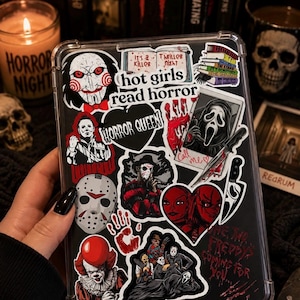 Puede incluir: Una funda transparente para tableta cubierta con pegatinas de terror, incluyendo el texto "hot girls read horror", "HORROR QUEEN" y "One, two, Freddy's coming for you". Las pegatinas muestran personajes icónicos como Billy, Ghostface, Jason Voorhees y Pennywise.