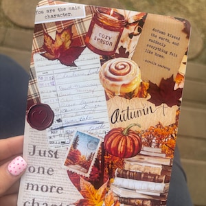 May include: A rectangular card featuring an autumn-themed collage with a candle, a cinnamon roll, a pumpkin, stacked books, and fall leaves. Text includes 'You are the main character', 'cozy season', 'Autumn kissed the earth', and 'Just one more chapter'.