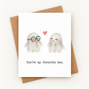 May include: A white greeting card featuring two watercolor ghosts with pink cheeks, one wearing black round glasses, separated by a small red heart. The text below reads, "You're my favorite boo." The card is placed on a brown envelope.