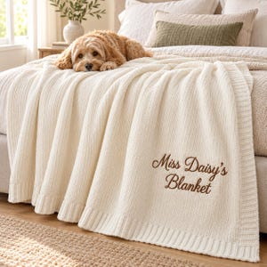 May include: A cream-coloured knitted throw blanket with a ribbed texture and a wide border. The text "Miss Daisy's Blanket" is embroidered in brown script on the lower right corner. A golden-brown dog is resting on top of the blanket.