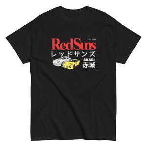 Peut inclure: Un t-shirt noir à manches courtes avec un imprimé graphique comprenant le texte rouge RedSuns, le texte blanc EST 1995 et des caractères japonais. Le motif inclut une voiture de sport blanche et une jaune, avec le texte AKAGI et des kanjis.