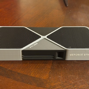May include: A black and silver graphics card featuring a dual-fan cooling system with a metallic X-shaped frame. The text RTX 5090 is printed on the central housing, and the text GEFORCE RTX is visible on the side of the cooling shroud.