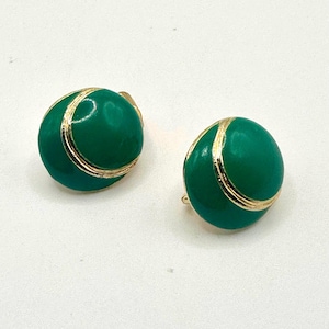 May include: A pair of round clip-on earrings featuring a smooth, glossy emerald green enamel finish. Each earring is accented with a thin, curved gold-tone metal line that sweeps across the surface, creating a distinct, polished, and reflective appearance.