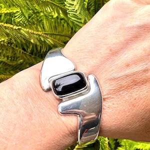 Peut inclure: Un bracelet manchette en argent poli orné d'une pierre noire rectangulaire et lisse au centre. La bande métallique présente un design large et asymétrique avec des bords incurvés qui se rétrécissent vers le poignet, offrant une finition brillante.