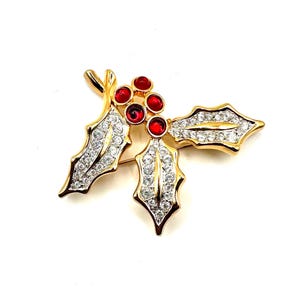 Op de afbeelding: Een goudkleurige metalen broche in de vorm van een hulsttakje, voorzien van drie ronde rode stenen die bessen voorstellen en drie puntige bladeren ingelegd met heldere, sprankelende strass-steentjes. De broche heeft een gepolijste afwerking en een speld.