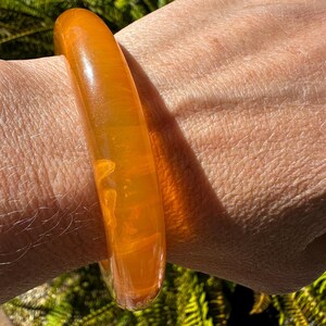 Puede incluir: Una pulsera tipo brazalete de color naranja translúcido y superficie lisa con un perfil redondeado y sutiles variaciones internas similares a nubes. El material tiene un acabado brillante que capta la luz, creando un aspecto cálido y radiante.