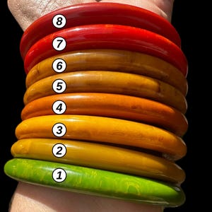 May include: A stack of eight smooth, rounded bangle bracelets in various colors, including green, yellow, orange, and red. Each bracelet is marked with a white circular label containing a black number from one to eight.