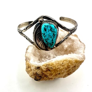 Puede incluir: Un brazalete de plata con una piedra turquesa central en forma de lágrima con vetas negras. La banda de metal está decorada con un motivo de hoja tallada en un lado y patrones geométricos grabados a lo largo de los brazos divididos.