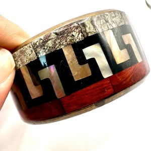 May include: A wide bangle bracelet featuring a multi-layered design. The top band consists of textured grey stone, followed by a central black inlay with a repeating iridescent mother-of-pearl Greek key pattern, and a bottom band of reddish-brown wood.