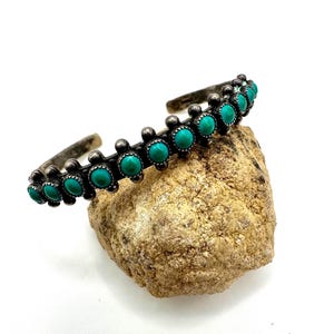 May include: A silver cuff bracelet featuring a row of small, round, vibrant turquoise stones set in individual bezels. The metal has a darkened, oxidised finish with decorative silver bead accents placed between each stone setting.