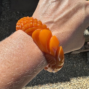 Op de afbeelding: Een dikke, doorschijnende oranje armband met een uitgesneden, gelaagd, bloembladachtig ontwerp dat om de pols valt. Het oppervlak van de armband heeft een gladde, gepolijste afwerking met diepe, ritmische groeven in een herhalend patroon.