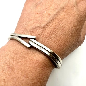 May include: A silver-toned metal cuff bracelet featuring a smooth, polished finish and a unique overlapping design where the two ends meet. The bracelet has a solid, slightly curved band structure that wraps around the wrist with a clean, metallic look.
