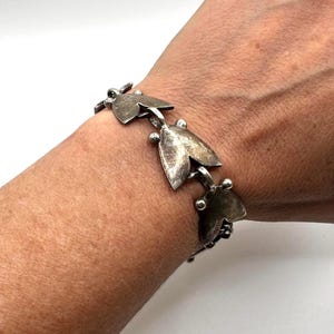 Könnte beinhalten: Ein Armband aus silberfarbenem Metall mit einer Reihe von sich wiederholenden blattförmigen Gliedern. Jedes Glied ist durch kleine kugelförmige Perlen und Metallringe verbunden, wodurch ein durchgehendes Kettendesign entsteht, das das Handgelenk umschließt.