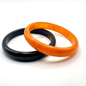 May include: Two smooth, circular bangle bracelets. One bangle is a solid, glossy black colour, while the other is a vibrant, translucent orange with a marbled, swirling texture. Both bracelets have a rounded, thick profile and a polished finish.