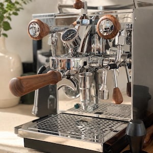 Könnte beinhalten: Eine Espressomaschine aus Chrom mit polierter Metalloberfläche, einem Manometer und Holzgriffen am Siebträger sowie an den Dampfreglern. Ein schwarzer Tamper mit der Aufschrift 'normcore' steht im Vordergrund auf der Oberfläche.
