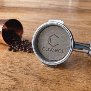 Puede incluir: Un portafiltro de espresso de acero inoxidable con mango de madera y un inserto de filtro de papel circular que presenta el texto coweet y un logotipo hexagonal, acompañado de una taza de metal martillado color cobre con granos de café.