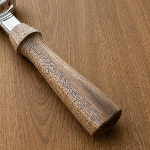 Puede incluir: Un mango de portafiltro de café hecho de madera marrón con un patrón de grano visible, unido a un componente de metal pulido de color plateado. El mango presenta una forma cilíndrica que se estrecha ligeramente hacia la unión metálica.