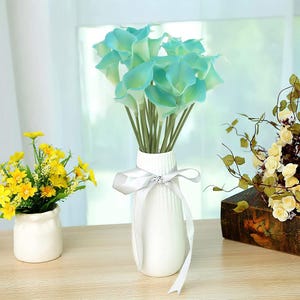 Könnte beinhalten: Ein Strauß künstlicher Calla-Lilien in Türkis- und Hellgrüntönen, arrangiert in einer hohen, weißen, vertikal gerippten Keramikvase. Die Vase ist mit einem einfachen weißen Band verziert, das zu einer Schleife um den schmalen Hals gebunden ist.