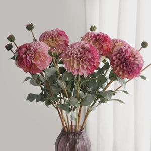 Puede incluir: Un ramo de flores de dalia artificiales con pétalos en capas en tonos rosa, magenta y crema. Los tallos son verdes con varias hojas y algunos pequeños capullos sin abrir, dispuestos en un jarrón de vidrio morado con relieve estriado.