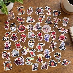 May include: A collection of assorted vinyl stickers featuring various animated characters from Helluva Boss, including individual portraits and group scenes. The stickers display vibrant colours and include a circular logo sticker with the text Helluva Boss.