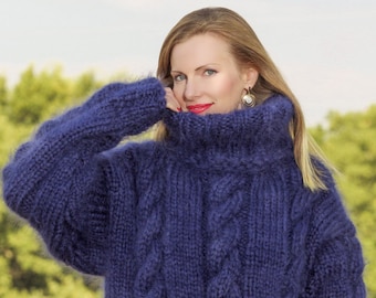May include: A navy blue, oversized cable-knit sweater with a high turtleneck. The sweater has a thick, textured appearance and is made of a soft, fluffy material. The image includes the text "SuperTanya" and "Hand crafted knitwear".