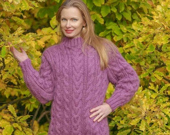 May include: A woman wearing a long, purple cable knit sweater with a high neck. She is standing in front of a background of fall foliage.