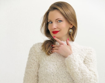 May include: Cream-colored, textured knit bodysuit with long sleeves and a high neckline. The bodysuit is from the brand SuperTanya, which is printed at the bottom of the image. The model is wearing gold earrings and red lipstick.