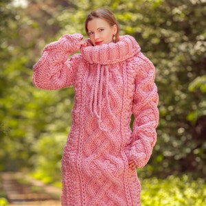 May include: A long, pink, chunky-knit sweater dress with a high, ribbed turtleneck and a drawstring. The dress features a cable-knit pattern and long sleeves. The model is standing on a path in a wooded area.