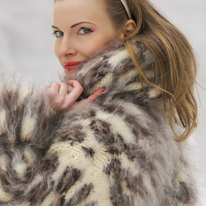 May include: A fluffy, brown and white knit sweater with a high collar. The sweater is made of mohair and has a unique, hand-crafted look. The text "SuperTanya® Hand crafted knitwear" is visible at the bottom of the image.