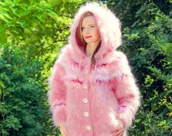 May include: A fluffy, light pink hooded coat with a button-up front. The coat features horizontal stripes of varying shades of pink and white. The model is wearing the coat outdoors, surrounded by green foliage.
