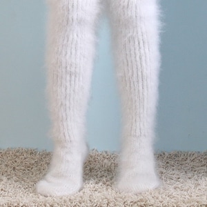 May include: White, fluffy, over-the-knee leg warmers. These leg warmers are made of a soft, fuzzy material and have a ribbed knit design. They are tied at the top with a white string. The leg warmers are shown against a light blue background.