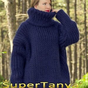 May include: A navy blue oversized turtleneck sweater made of soft yarn. The sweater is hand-knitted and has a loose, comfortable fit. The sweater is photographed against a blurred background of trees.