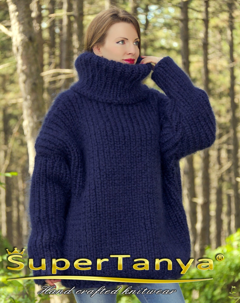 May include: A navy blue oversized turtleneck sweater made of soft yarn. The sweater is hand-knitted and has a loose, comfortable fit. The sweater is photographed against a blurred background of trees.