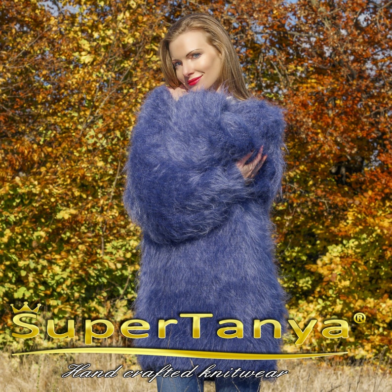 May include: A woman wearing a long, fluffy, periwinkle blue knit sweater. The sweater has long sleeves and a soft, textured appearance. The background features autumn foliage with golden and brown leaves. The text "SuperTanya" and "Hand crafted knitwear" are visible.