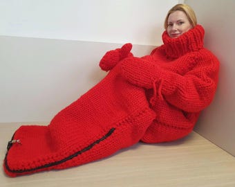 May include: A large, bright red knitted wearable blanket with a high turtleneck collar and attached mittens. The blanket has a zipper closure at the foot and is designed for full-body coverage. The item is made of thick, chunky knit material.
