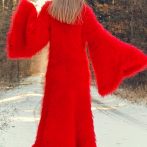 May include: A long, vibrant red, fuzzy coat with wide bell sleeves. The coat is ankle-length and appears to be made of a soft, plush material. The person is standing on a snow-covered path in a forest.
