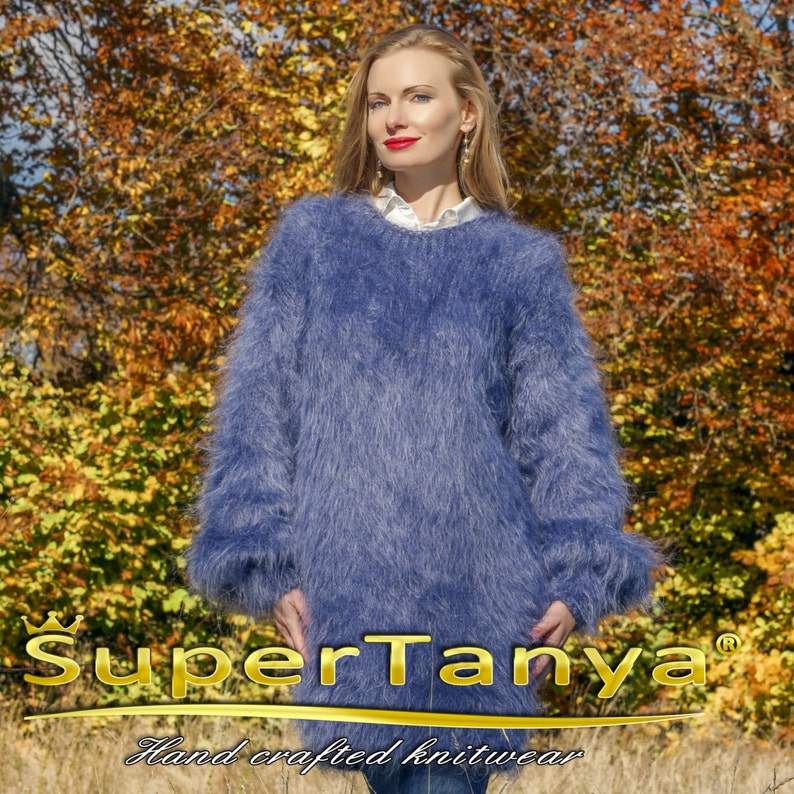 May include: A long-sleeved, blue knit sweater with a fluffy texture. The sweater has a crew neck and long sleeves. The text "SuperTanya" is displayed in gold, along with "Hand crafted knitwear". The background features autumn foliage.
