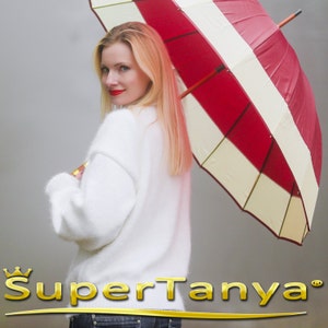 May include: A woman with long blonde hair is holding a red and white umbrella. She is wearing a white sweater and jeans. The umbrella has a wooden handle.